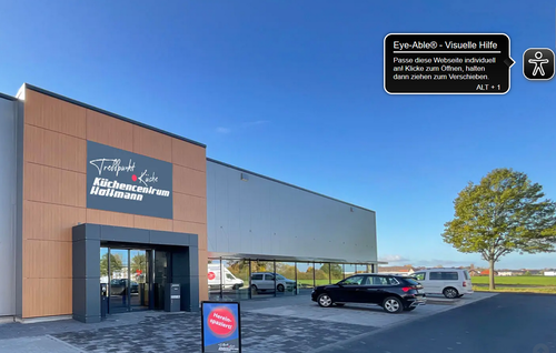 Moderne Außenansicht des Küchenzentrum Holtmann mit großer Glasfront, parkenden Autos und freistehendem Baum auf dem Parkplatz an einem klaren Sonnentag