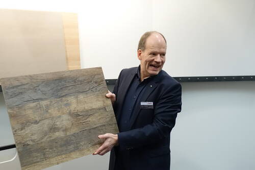 Mann im Anzug präsentiert lächelnd eine große Holzplatte mit rustikaler Maserung vor einer hellen Wand.