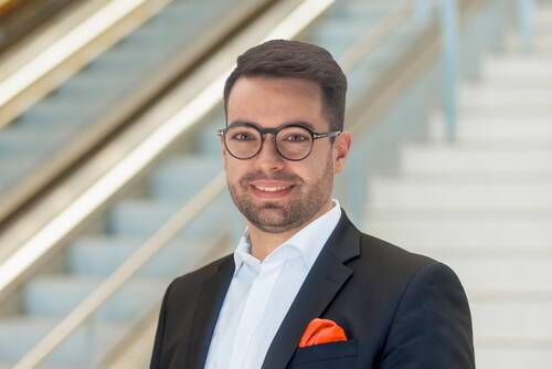 Junger Mann im Anzug mit orangenem Einstecktuch und runder Brille lächelt vor einer modernen Treppenanlage in einem Bürogebäude