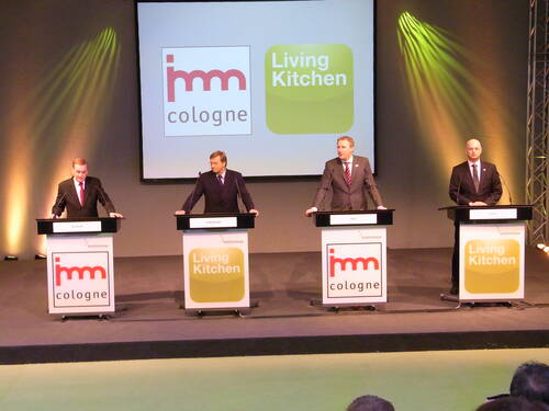 Vier Geschäftsleute in Anzügen stehen an Rednerpulten auf einer Bühne der Koelnmesse, während hinter ihnen groß die Logos von „imm cologne“ und „Living Kitchen“ projiziert werden.