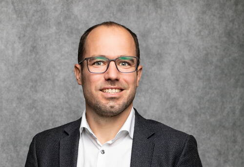 Mann mittleren Alters mit Brille, weißem Hemd und dunklem Sakko lächelt vor neutral grauem Hintergrund in die Kamera