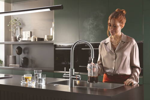 Frau in moderner Küche füllt mit kochend heißem Wasser aus einem designorientierten Wasserhahn eine French Press auf einer eleganten Kücheninsel.