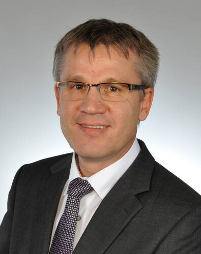 Mittelalterlicher Mann mit Brille im grauen Anzug und gemusterter Krawatte lächelt vor neutral grauem Hintergrund in die Kamera.