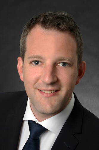 Junger Mann im Anzug und Krawatte lächelt direkt in die Kamera vor dunklem, neutralem Hintergrund.