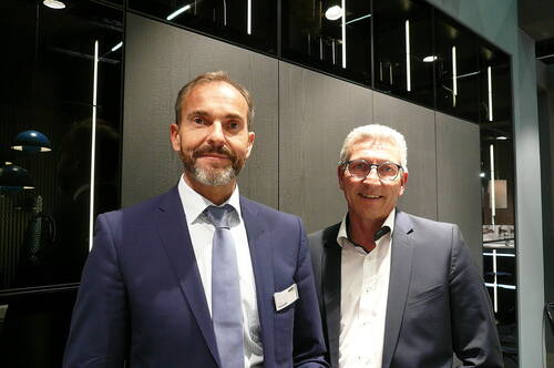 Zwei lächelnde Geschäftsmänner in Anzügen stehen in einem modernen, dunkel gestalteten Büro- oder Messeraum mit Glasfronten und vertikaler Beleuchtung im Hintergrund.