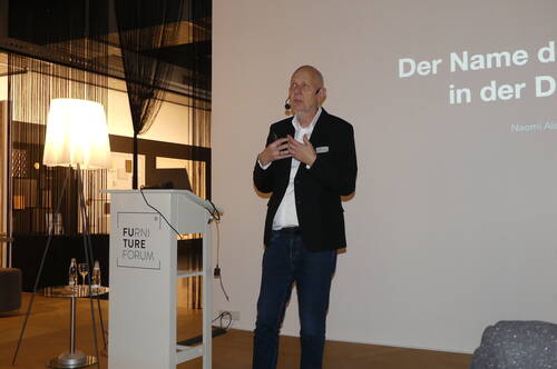 Redner im schwarzen Sakko hält mit Headset-Mikrofon einen Vortrag am Rednerpult des Furniture Forums vor projizierter Präsentationsfolie.