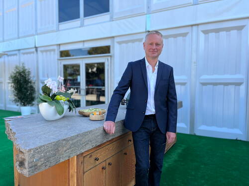 Geschäftsmann im dunklen Anzug steht an einer Stein-Theke mit Orchideendeko vor einem modernen weißen Gebäude mit Glas­eingang.