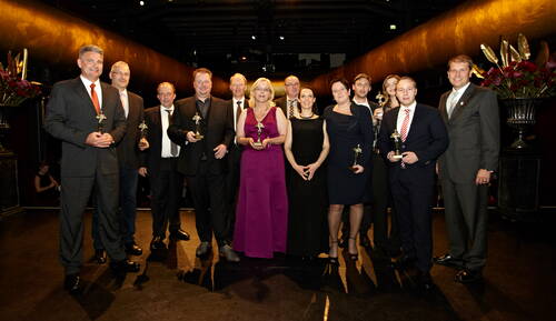 Gruppe elegant gekleideter Preisträger steht in einem festlich beleuchteten Saal und hält goldene Trophäen in den Händen