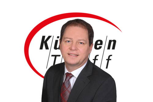 Geschäftsmann im dunklen Anzug mit karierter Krawatte lächelt vor einem weißen Hintergrund mit rotem Bogen und dem Schriftzug „Küchen Treff“.