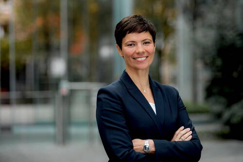 Selbstbewusste Geschäftsfrau mit kurzhaarigem Haarschnitt und dunkelblauem Blazer steht mit verschränkten Armen lächelnd vor einem modernen Glasgebäude