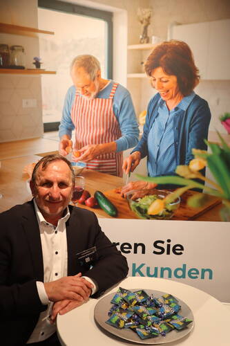 Älterer Mann im Anzug sitzt lächelnd an einem Messetisch mit Schüssel voller Werbesüßigkeiten vor einem großen Plakat, das ein älteres Paar beim gemeinsamen Gemüseschneiden in einer hellen Küche zeigt