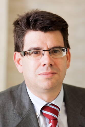 Mittelalterlicher Mann mit kurzen dunklen Haaren, Brille und Anzug mit rot‑blau gestreifter Krawatte blickt ruhig in die Kamera vor neutralem Hintergrund.