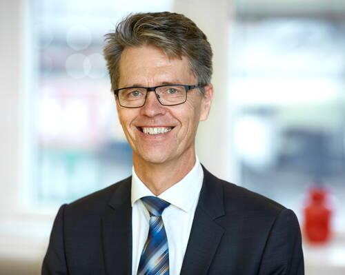 Lächelnder Mann im Anzug mit blau gestreifter Krawatte und Brille steht in einem hellen Büro mit unscharfem Hintergrund