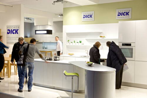 Besucher betrachten eine moderne, weiß eingerichtete Einbauküche an einem Messestand von „Küchen Dick“, während ein Mitarbeiter im Hintergrund Auskunft gibt.