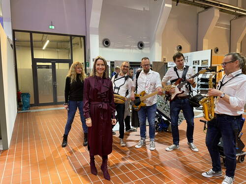 Elegante Frau in bordeauxrotem Kleid steht lächelnd vor einer Band aus fünf Musikern mit Gitarren, Bass und Saxofon in einer modernen Halle mit roten Bodenfliesen.