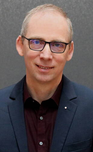 Mittelalterlicher Mann mit Brille im dunkelblauen Sakko und bordeauxrotem Hemd lächelt vor neutral grauem Hintergrund
