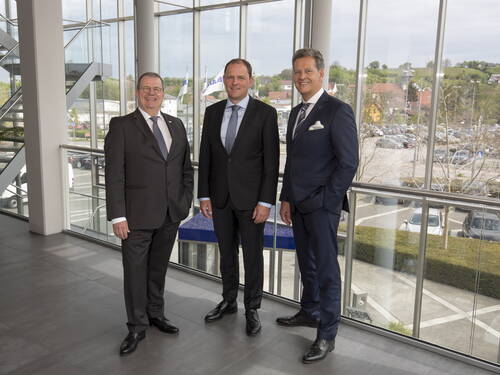 Drei geschäftsmänner in Anzügen posieren lächelnd in einem modernen, verglasten Bürogebäude mit Blick auf den Parkplatz und die umliegende Stadt