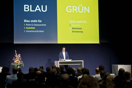 Redner erklärt vor Publikum eine Präsentation, die die Bedeutung der Farben Blau für Ruhe, Stabilität und Verantwortung sowie Grün für Finanzen, Wachstum und Erneuerung gegenüberstellt