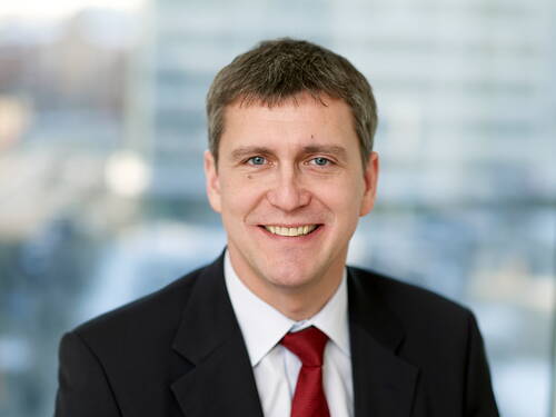 Lächelnder Mann im Anzug mit roter Krawatte steht vor unscharfem Hintergrund in einem modernen Bürogebäude