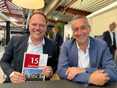 Zwei lachende Männer im Anzug sitzen an einem Tisch in einer modernen Ausstellungshalle, einer hält eine Zeitschrift mit der Titelzeile „15 küchennews“ in die Kamera.
