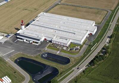 Großes, modernes Industriegebäude mit hellen Flachdächern, Laderampen und Parkflächen, umgeben von Feldern, Straßen und mehreren künstlichen Wasserbecken in der Vordergrundzone