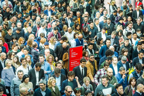 Dicht gedrängte Menschenmenge strömt durch die Eingangshalle der Messe Salone del Mobile in Mailand, während ein rotes Wegweiserschild zur Rezeption und zum Pressebereich führt.