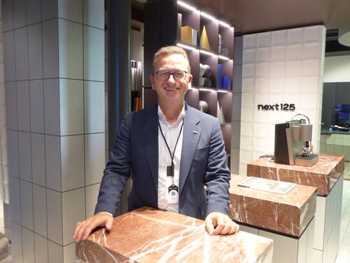 Lächelnder Mann im Anzug steht an einem braunen Marmortresen in einem modernen Showroom mit Designregalen und Kaffeemaschine der Marke „next125“.