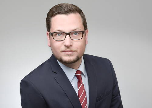 Junger Mann mit Brille im dunklen Anzug und rot-weiß gestreifter Krawatte blickt ernst in die Kamera vor neutralem Hintergrund