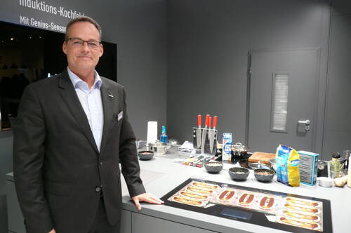 Geschäftsmann im Anzug präsentiert modernes Induktionskochfeld in Showküche mit vorbereiteten Speisen, Messern und Zutaten auf der Arbeitsfläche
