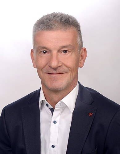 Mittelalterlicher Mann mit kurzem grauem Haar im dunklen Anzug und weißem Hemd, der freundlich in die Kamera lächelt vor neutralem, hellem Hintergrund.