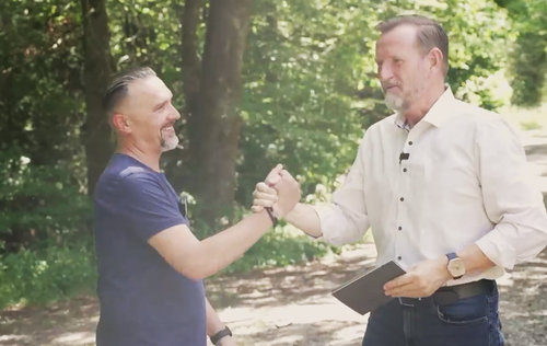Zwei Männer begrüßen sich lachend mit Handschlag im sonnigen Waldweg, einer hält ein Notizbuch in der Hand.