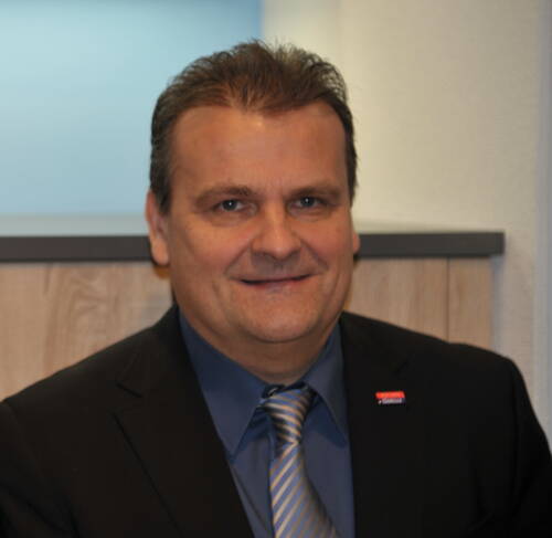 Mittelalterlicher Mann im Business-Anzug mit blauer Krawatte lächelt leicht in einem modernen Büroambiente.