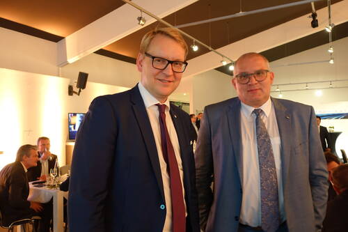 Zwei Männer in Anzügen und Krawatten stehen in einem hell erleuchteten Veranstaltungsraum und blicken lächelnd in die Kamera, während im Hintergrund Gäste an Tischen sitzen und sich unterhalten.