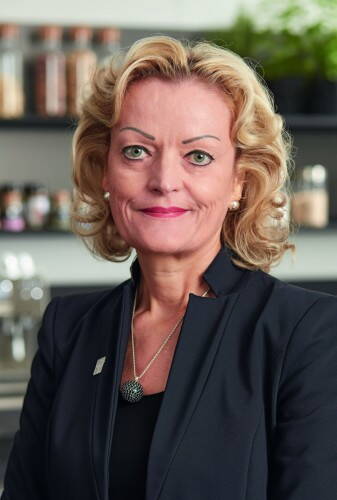 Elegant gekleidete Geschäftsfrau mit blond gelockten Haaren lächelt selbstbewusst in moderner Büroumgebung in die Kamera.