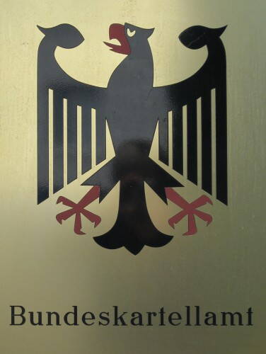 Schwarzer Bundesadler mit roten Krallen und Schnabel über der Aufschrift „Bundeskartellamt“ auf einer goldenen Metalltafel