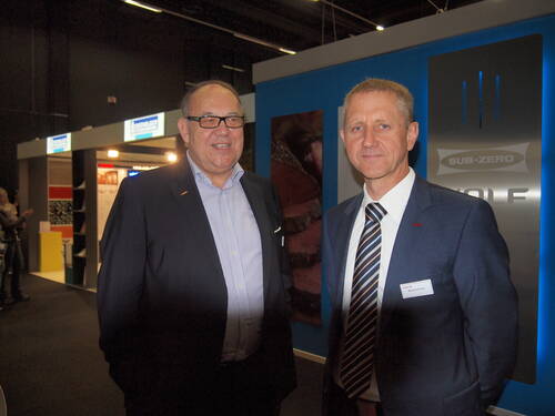 Zwei Geschäftsleute im Anzug lächeln in einer Messehalle vor einem Messestand mit blauem Hintergrund und „Sub-Zero“-Beschriftung in die Kamera