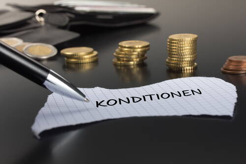 Kugelschreiber zeigt auf Zettel mit dem Wort „Konditionen“, im Hintergrund gestapelte Euromünzen und Geldbörse auf dunklem Tisch