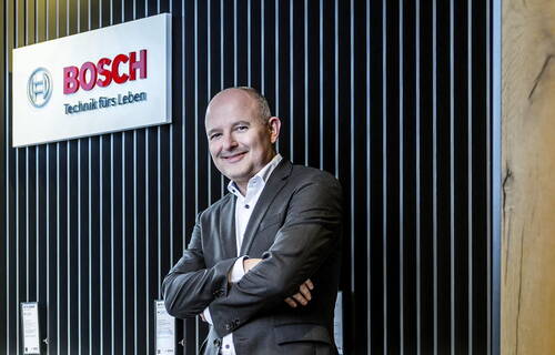 Geschäftsmann im Anzug mit verschränkten Armen steht lächelnd vor einer schwarzen Lamellenwand neben einem großen Bosch-Firmenlogo.