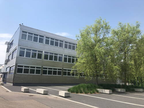 Modernes Bürogebäude mit metallisch schimmernder Fassade und großen Fenstern, eingerahmt von jungen Bäumen und leeren Parkplätzen an einem sonnigen Tag