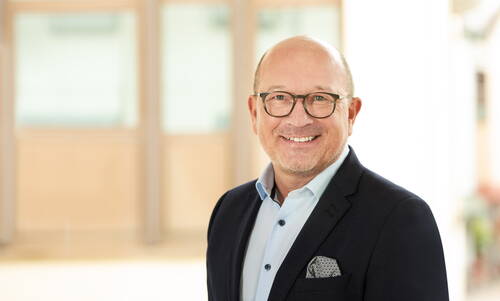 Älterer Mann mit Brille und Sakko lächelt freundlich in die Kamera vor einem hellen, unscharfen Bürohintergrund.