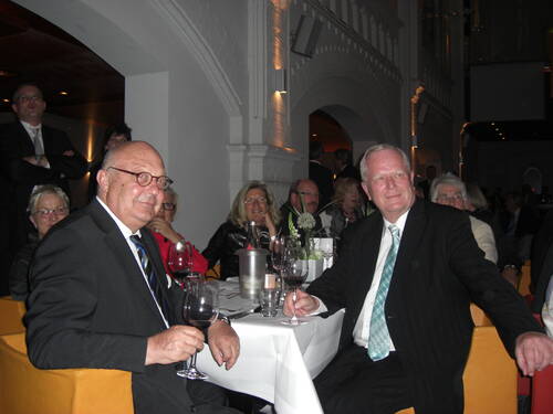 Ältere festlich gekleidete Gäste sitzen in einem stimmungsvoll beleuchteten Saal an einem weiß gedeckten Tisch, halten Weingläser in der Hand und blicken lächelnd in Richtung Kamera.