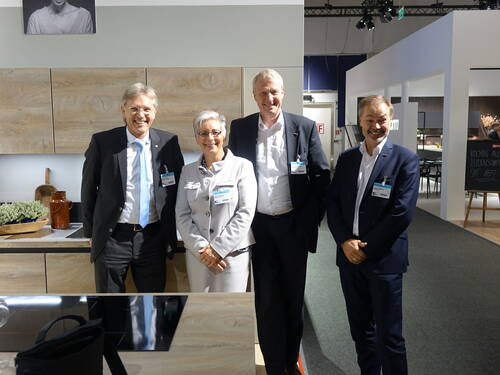 Vier Geschäftsleute in Anzug und Kostüm lächeln nebeneinander in einer modernen Küchenausstellung mit Holzmöbeln und Messeschildern im Hintergrund