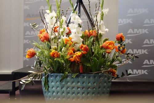 Üppiges Blumenarrangement aus orangefarbenen Tulpen und weißen Ritterspornblüten in einer großen blauen Keramikvase vor einer Bühne mit AMK-Logo im Hintergrund