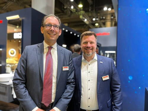 Zwei lächelnde Messemitarbeiter in Anzügen stehen nebeneinander an einem modernen, hell beleuchteten Messestand.
