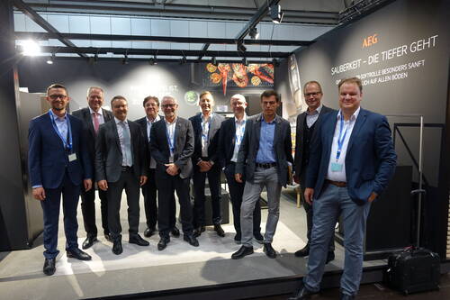 Gruppe von Geschäftsleuten in Anzügen posiert lächelnd vor einem AEG-Messestand mit Slogan zur Sauberkeit und Ausstellungsfläche für Haushaltsgeräte