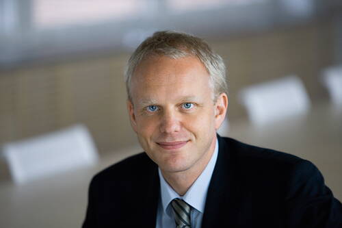 Blonder Geschäftsmann im Anzug lächelt in einem modernen, hellen Konferenzraum direkt in die Kamera