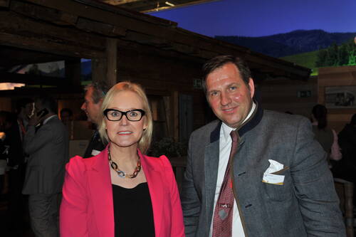 Elegant gekleidete Frau im pinken Blazer und Mann im traditionellen grauen Janker lächeln gemeinsam in einer rustikalen Eventlocation mit Holzwänden und Bergpanorama im Hintergrund