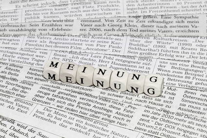 Holzwürfel mit dem Wort „MEINUNG“ liegen auf einer aufgeschlagenen Zeitungsseite voller Fließtext