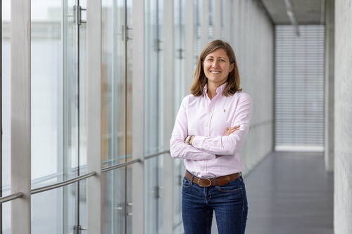 Junge Frau mit verschränkten Armen in rosa Hemd und Jeans steht lächelnd in einem modernen Büroflur mit Glasfronten.