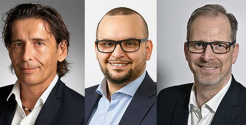 Drei lächelnde Männer in Businesskleidung stehen nebeneinander vor neutralem Hintergrund in einem professionellen Porträt.